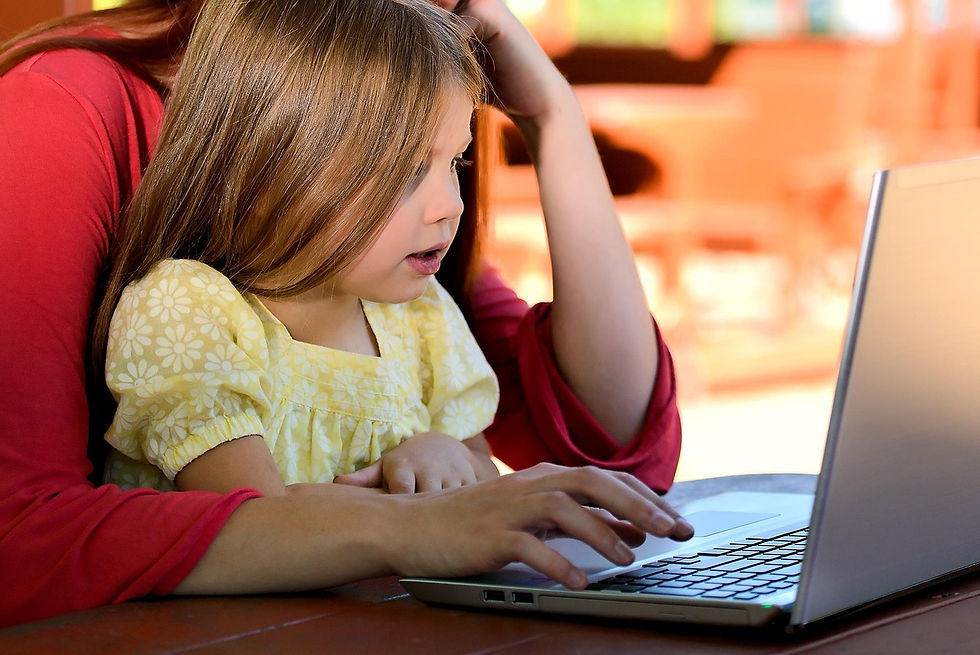 A child in a yellow dress uses a laptop, assisted by an adult in red. They're focused indoors, with warm lighting.