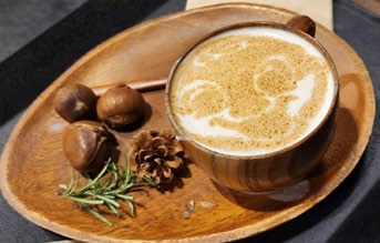 Latte art with a smiling face on a wooden tray, accompanied by chestnuts, rosemary, and a pine cone, creating a cozy autumn vibe.
