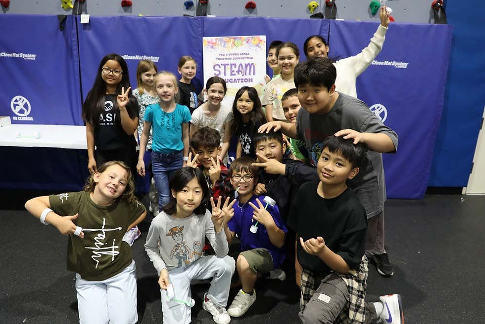 Group of smiling children posing in a gym, making peace signs. "STEAM" poster in background. Blue mats and colorful holds on walls.