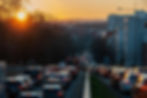 Traffic jam at sunset, cars and a bus on a busy road. Sun sets over buildings and trees, casting warm light on the scene. Mood: busy.