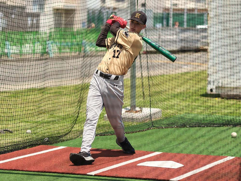 Baseball player in beige jersey swings a bat in a practice cage on green turf, surrounded by netting, with focus and determination.