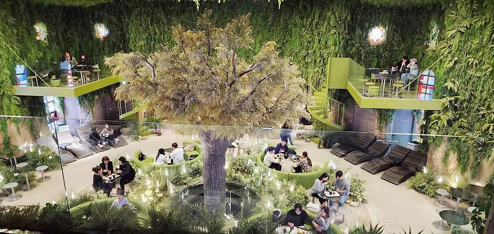 People sitting and dining in a lush, indoor garden café with a large artificial tree center, surrounded by greenery and soft lighting.