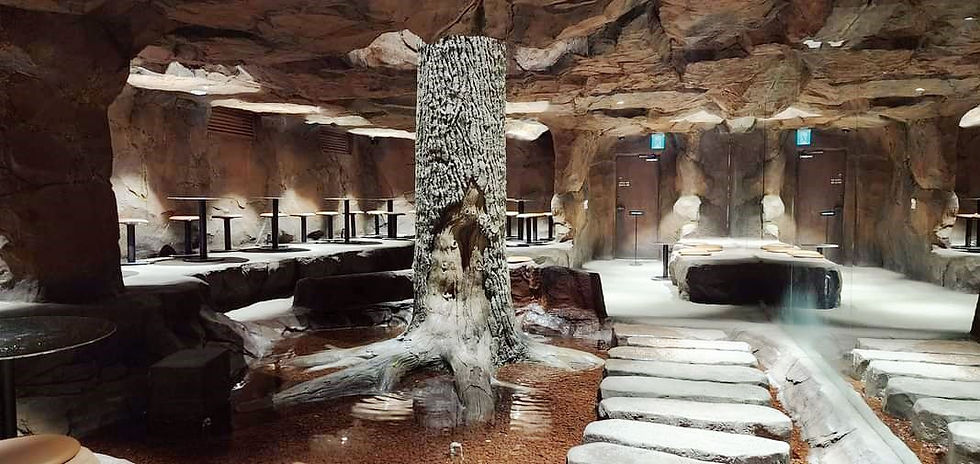 Cave-themed restaurant interior with stone walkways, a central tree, and round tables. Warm lighting creates a cozy atmosphere.