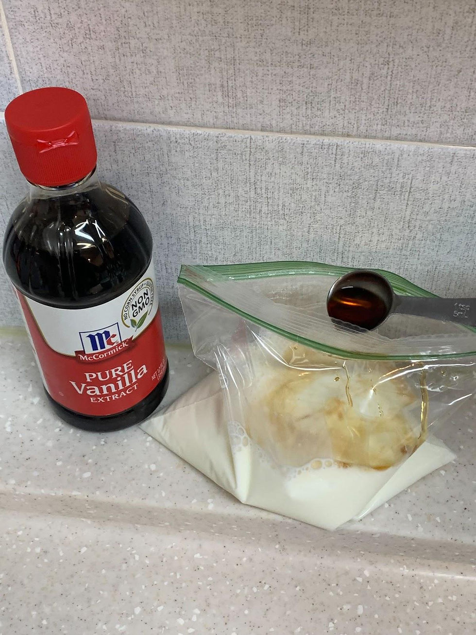 Vanilla extract bottle with red cap beside a sealed bag of milk and vanilla on a speckled countertop, set against a light tiled background.