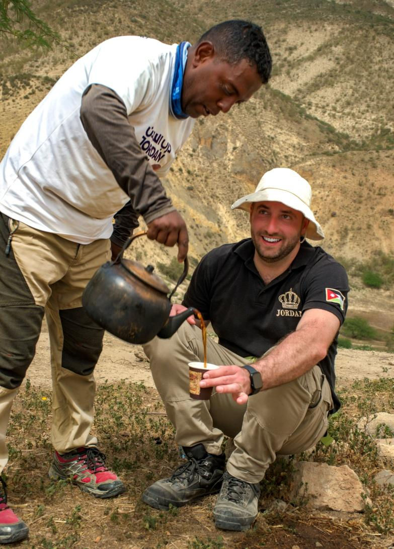 Two men outdoors, one pouring tea from a kettle into a cup. Arid landscape in the background. The mood is friendly and relaxed.