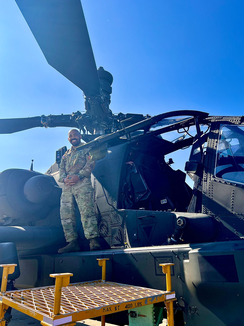 Smiling soldier in camo stands on helicopter wing under clear blue sky. Yellow platform labeled "MAX WT 400 LBS" is below.