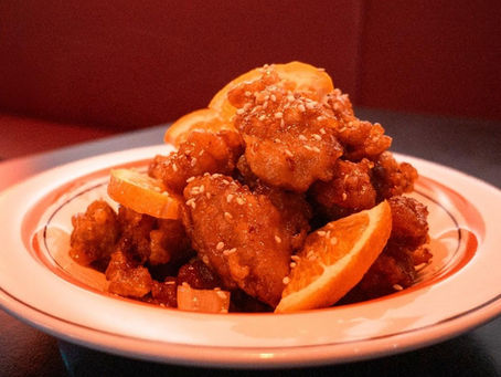 Plate of orange chicken with sesame seeds and orange slices, set on a restaurant table. Warm lighting creates a cozy ambiance.