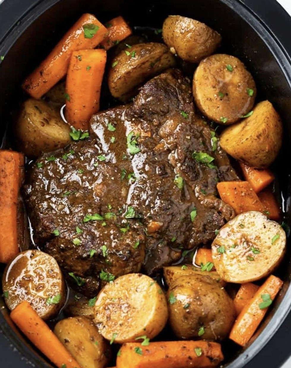 A pot roast with glazed carrots and potatoes in a black slow cooker, garnished with parsley, creating a warm, hearty meal vibe.
