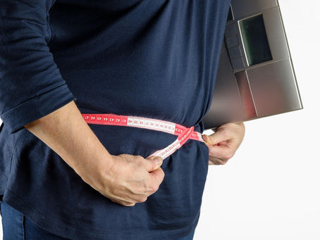 Person in a blue shirt measures waist with a red tape, holding a black scale. White background. Mood appears focused.