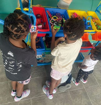 Three children stand by colorful toy bins, selecting toys. They wear casual clothes and sneakers on a tiled floor, creating a playful mood.