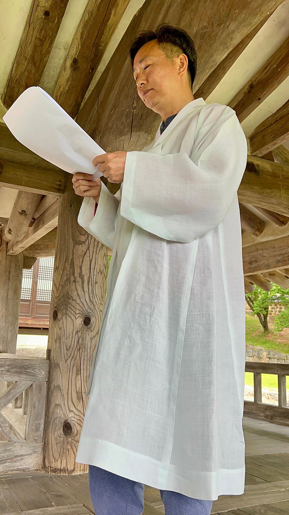 Man in white traditional clothing reads papers under a wooden pavilion. Greenery visible in the background. Serene atmosphere.