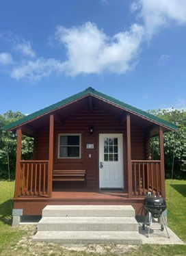 A small wooden cabin with a green roof sits under a blue sky. It has a porch with a bench, steps leading up, and a grill beside it.