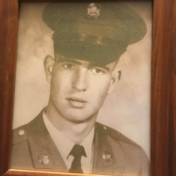 Black-and-white portrait of a young man in military uniform and hat, set in a wooden frame. The expression is serious and formal.