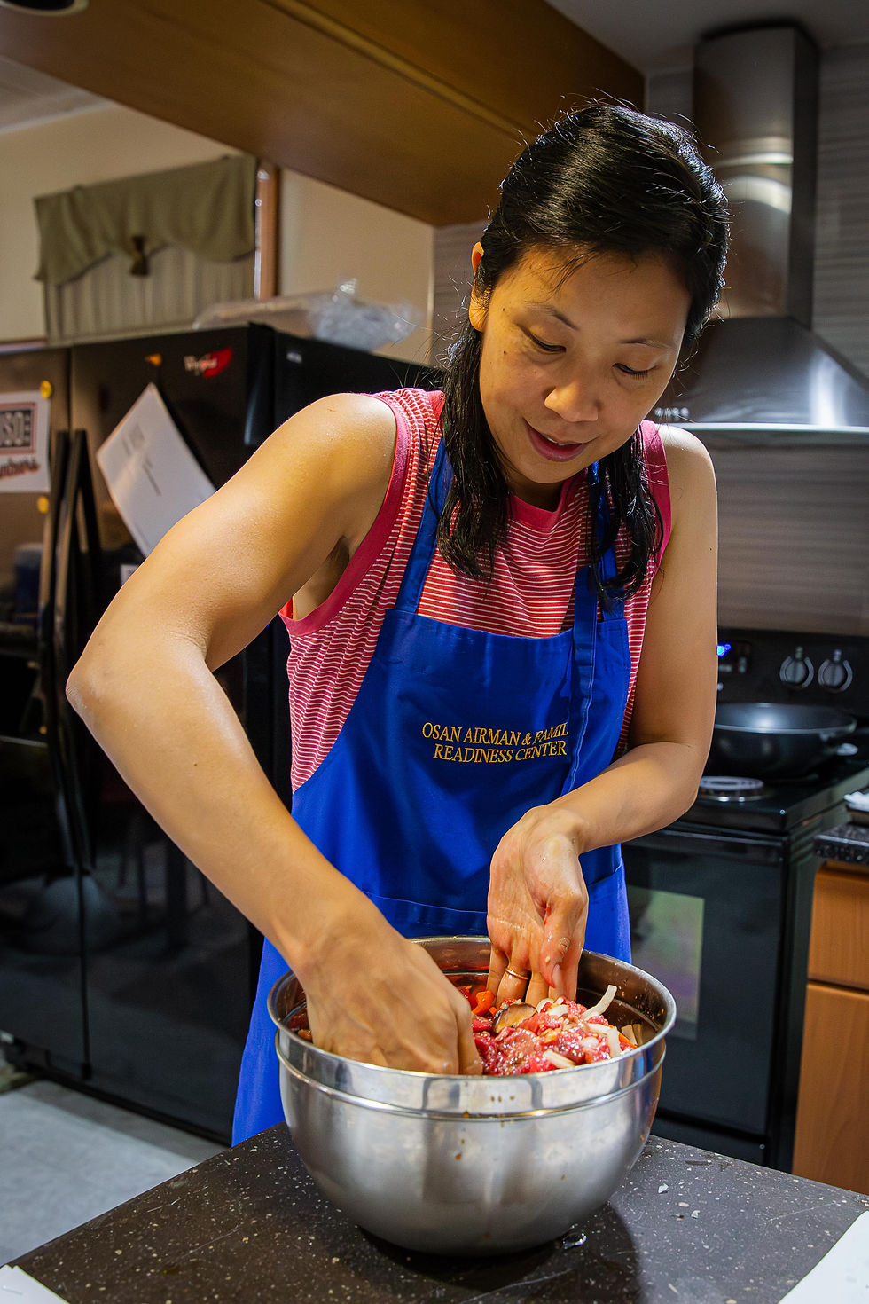 Woman in red and blue apron mixes ingredients in a steel bowl in a kitchen. Background has a stove and fridge. Text on apron visible.