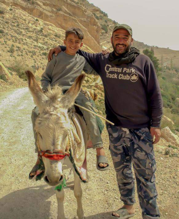 Man stands smiling beside a boy riding a donkey on a dirt path. Boy wears gray; man wears a "Central Park Track Club" sweatshirt. Rugged hills in background.
