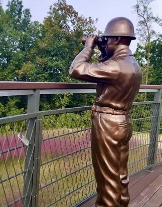 Bronze statue of a man with binoculars on a deck, surrounded by trees. Overcast sky, relaxed pose, gaze directed at the horizon.
