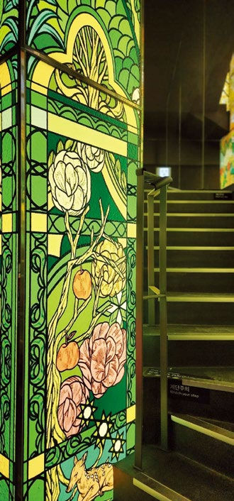 Stained glass panel with floral and tree designs in green and yellow tones next to a staircase. A warm, artistic atmosphere prevails.