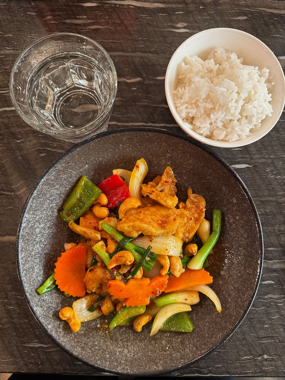Plate of stir-fry with vegetables and cashews, served with a bowl of white rice and a glass of water on a dark textured table.