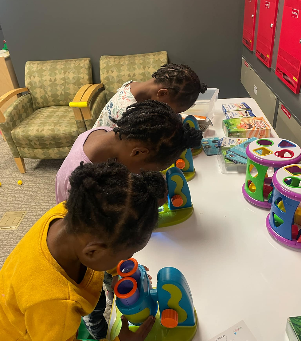 Three children look into toy microscopes on a table in a playroom. Bright colors and playful mood with patterned chairs in the background.