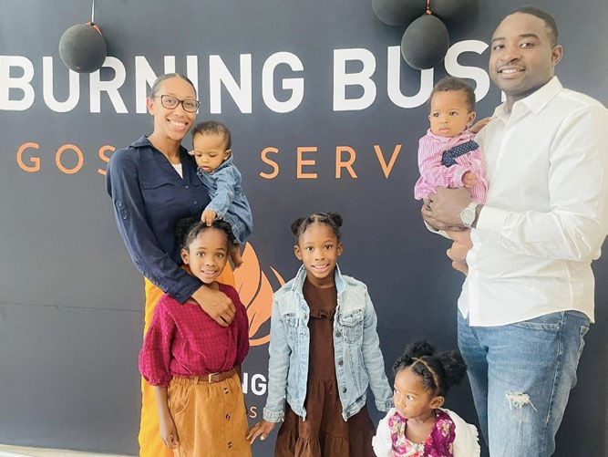 Family of six smiling in front of a "BURNING BUSH SERVICES" banner. Bright attire, happy mood, and black balloons in the background.