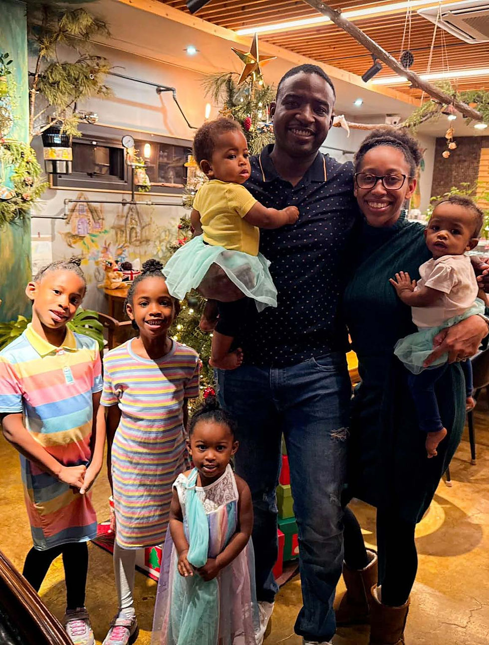 Family of six poses happily by a decorated Christmas tree. Children wear colorful outfits. Warm indoor setting with festive decor.