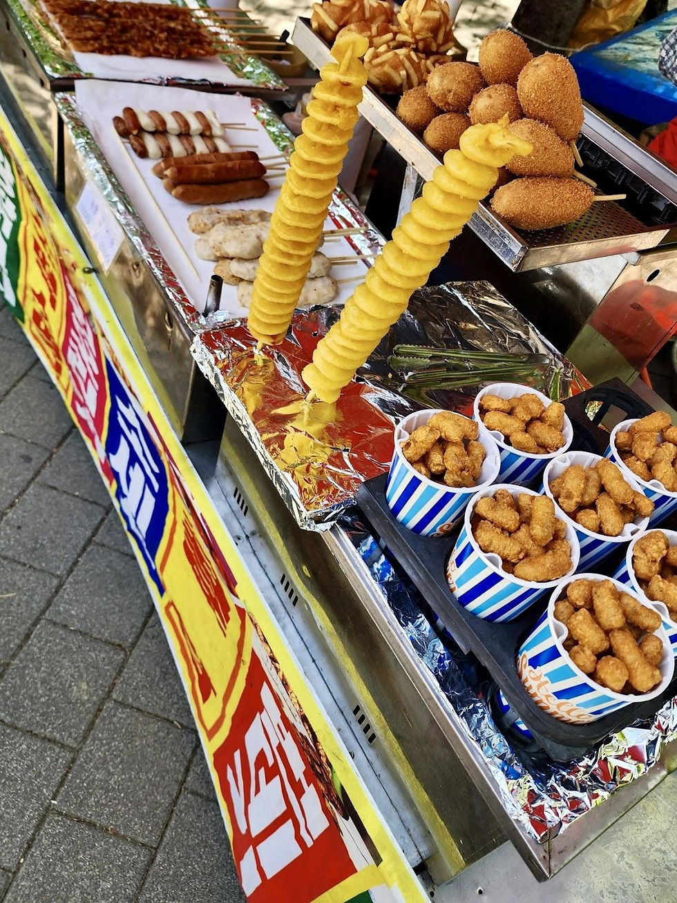 Street food stall displaying skewered potatoes, fried snacks, and sausages on foil trays. Bright banners and striped cups add color.