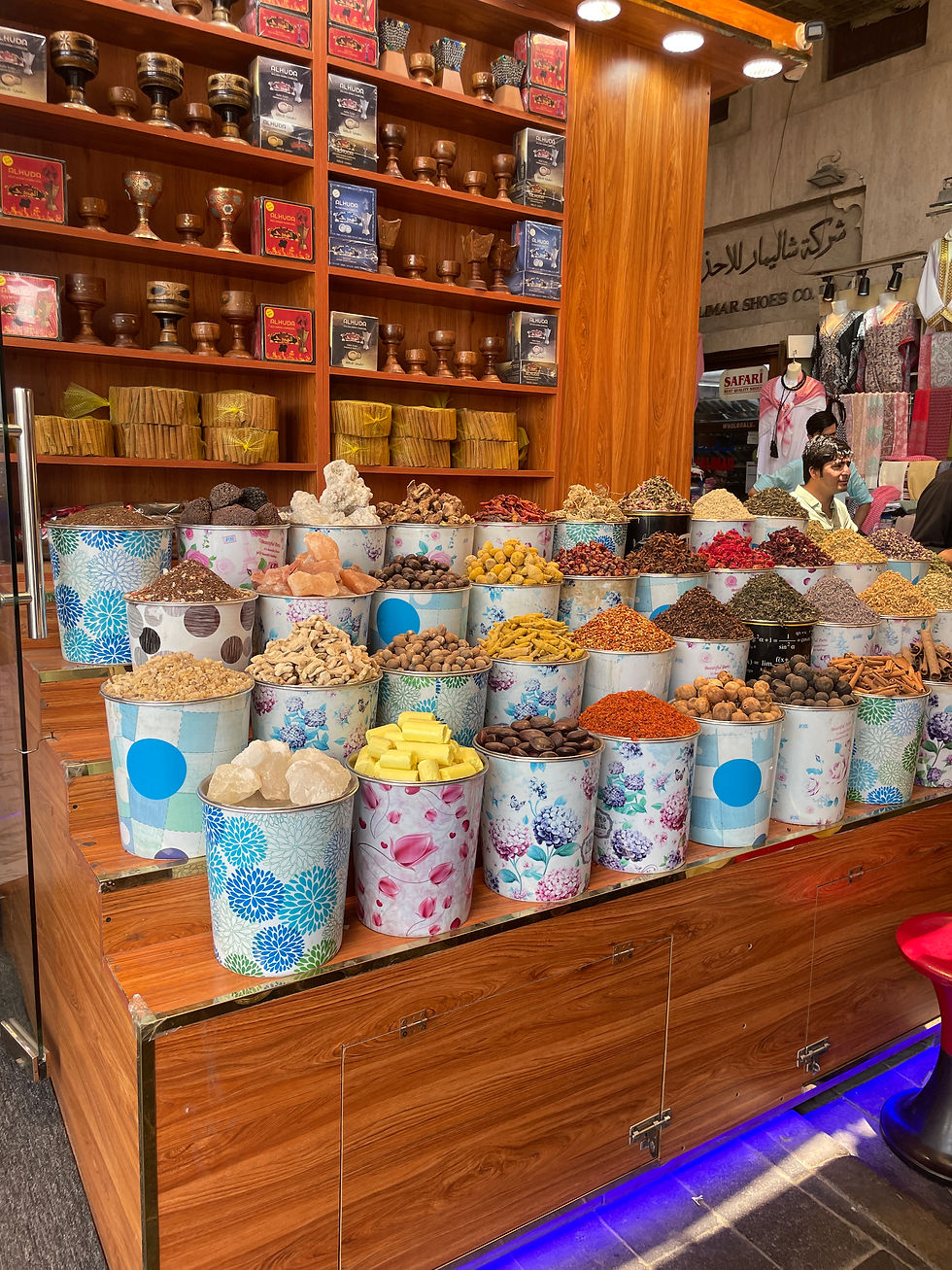 Colorful spice shop with floral buckets filled with various spices. Wooden shelves display goods. Arabic text and mannequins in the background.