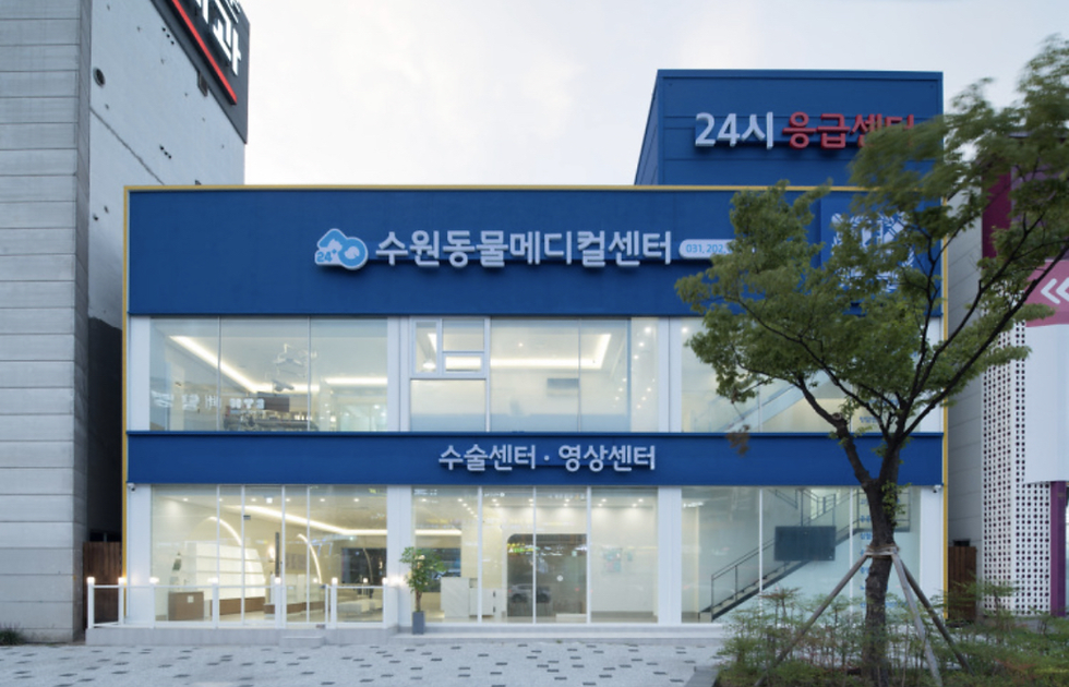 Blue building with Korean text, "24/7 Emergency," large windows, modern interior. Tree in foreground, overcast sky.