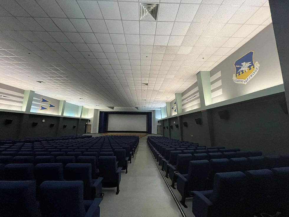 Empty theater with rows of blue seats, a large screen at the front. Walls have military insignia and text "Leading the Charge."