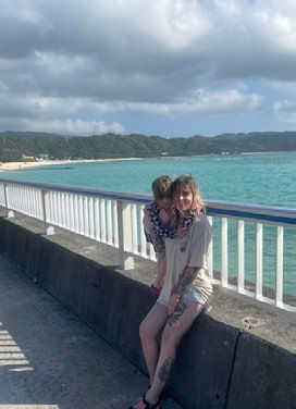A woman and child sit on a pier railing by a turquoise sea under cloudy skies. Mountains are in the background. The mood is peaceful.
