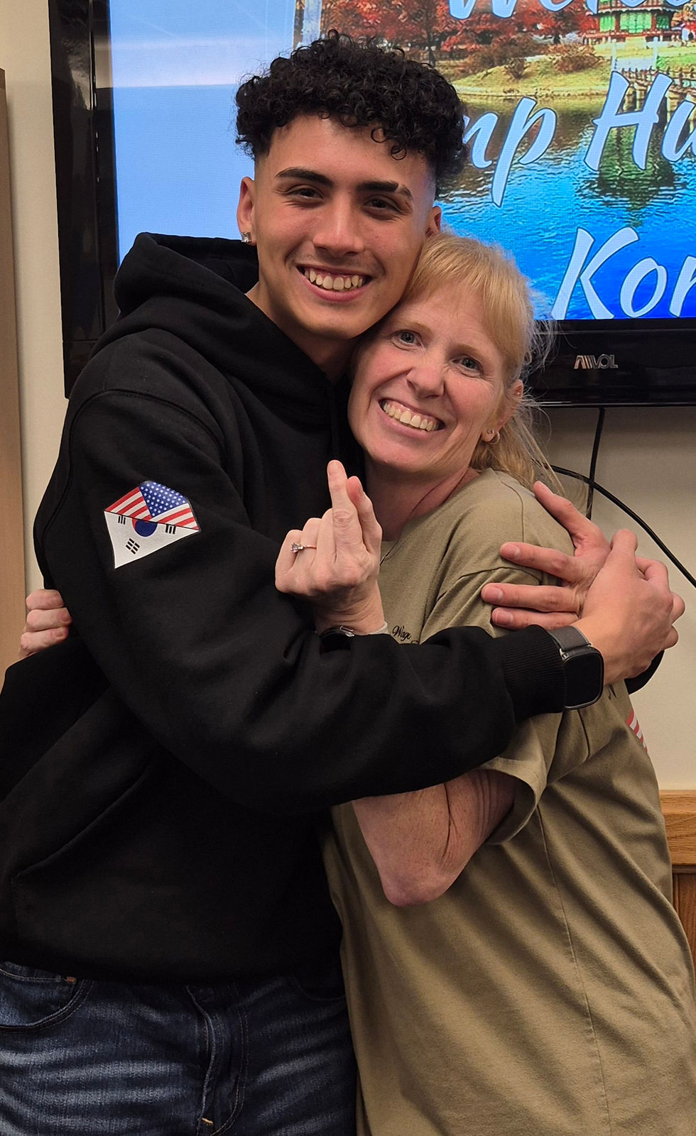 A smiling person in a black hoodie hugs another smiling person in a tan shirt. Text on the screen in the background reads "Camp Humphreys Korea."