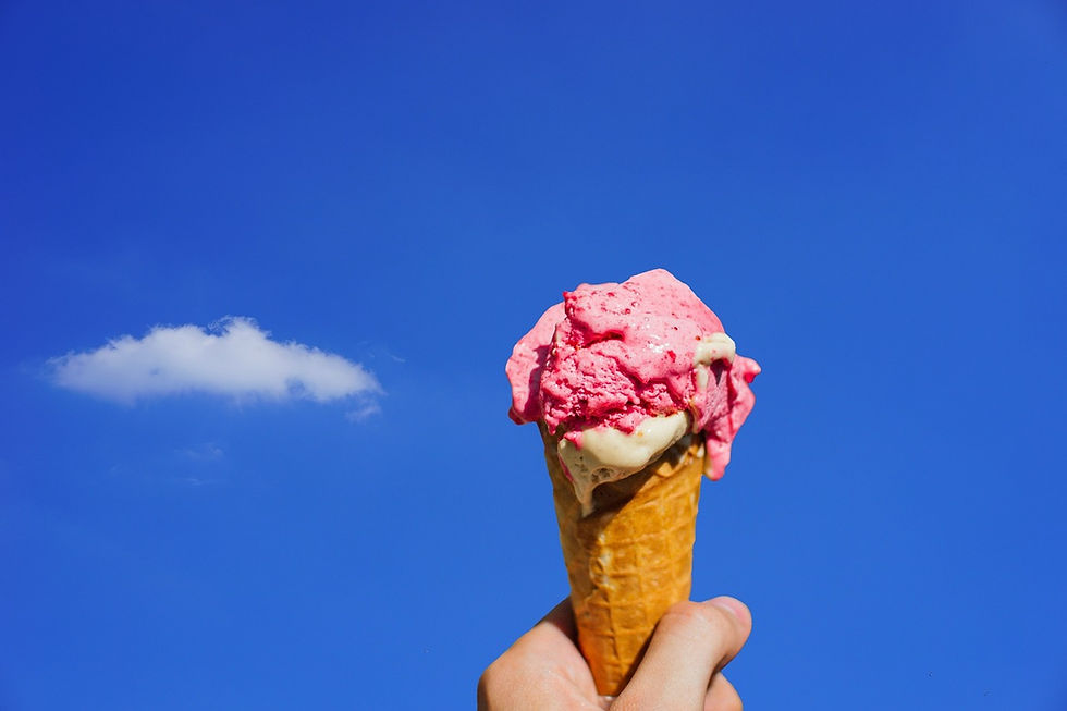 Hand holding ice cream cone with pink and white scoops against a clear blue sky, small cloud in background. Bright, summery feel.