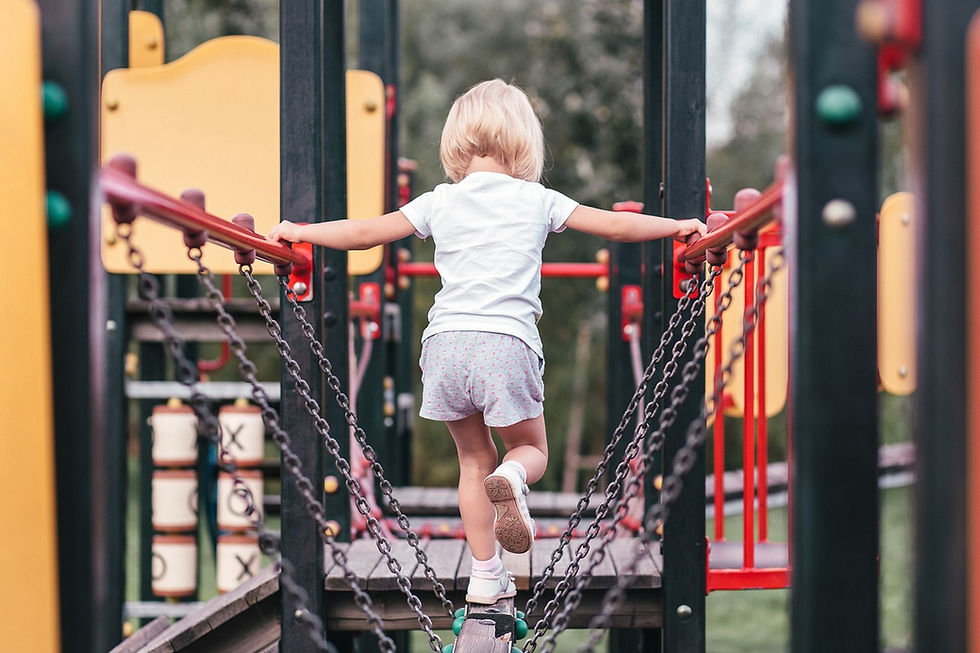 Child in white and gray crossing a chain bridge at a playground. Bright red and yellow structures in background, creating a playful scene.