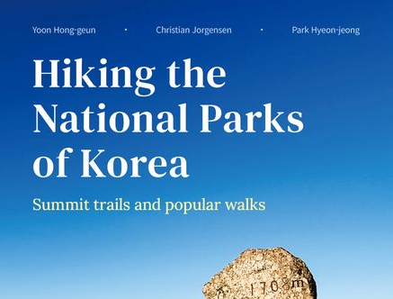 Stone marker with red Korean text on a mountain summit under a clear blue sky. Text: "Hiking the National Parks of Korea."