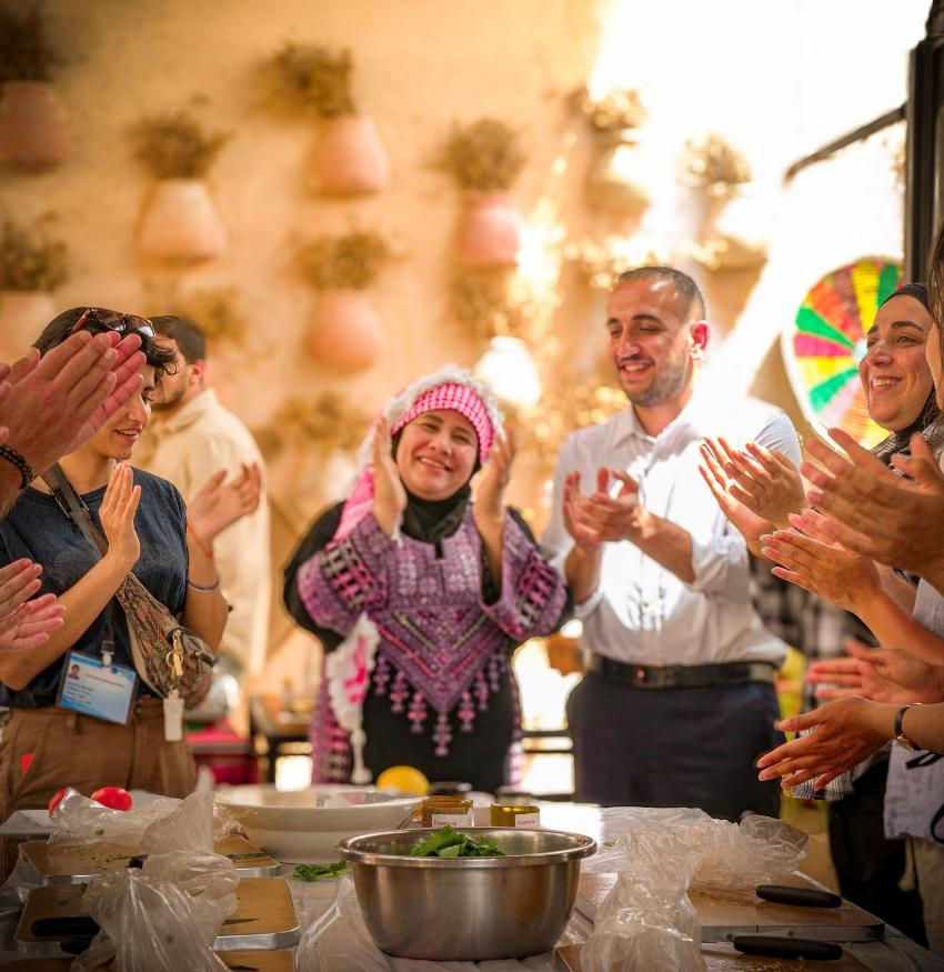 People wearing traditional attire clap joyfully around a table with bowls. Warm setting with hanging pots and natural light.