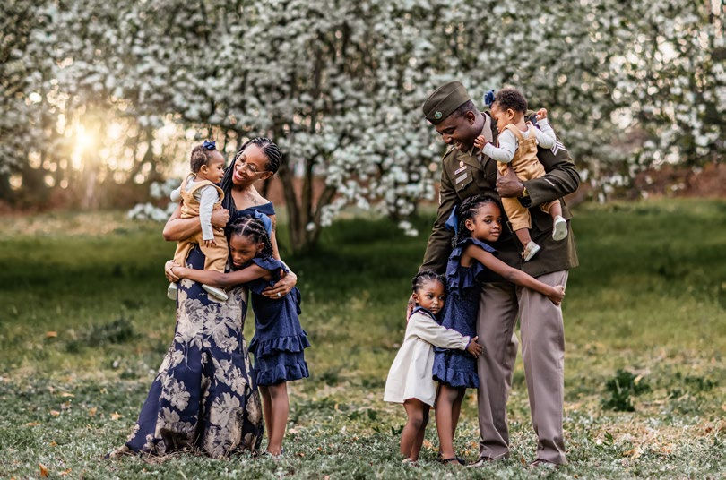 Family of six in a park, parents holding infants, three kids hugging them. Blossom trees in background, creating a warm, loving moment.