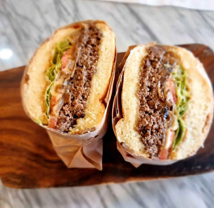 Two stacked burger halves on a wooden board, filled with lettuce, tomatoes, and beef patty, wrapped in brown paper. Marble surface background.