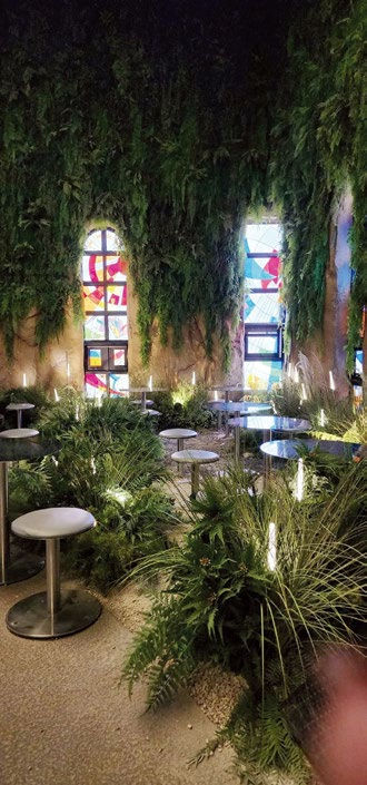 Indoor garden with metal stools and green plants under vibrant stained glass windows. Soft lighting creates a tranquil, natural ambiance.