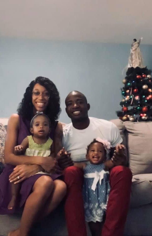 A family of four smiles on a sofa with a decorated Christmas tree in the background. The room has a cozy, cheerful atmosphere.