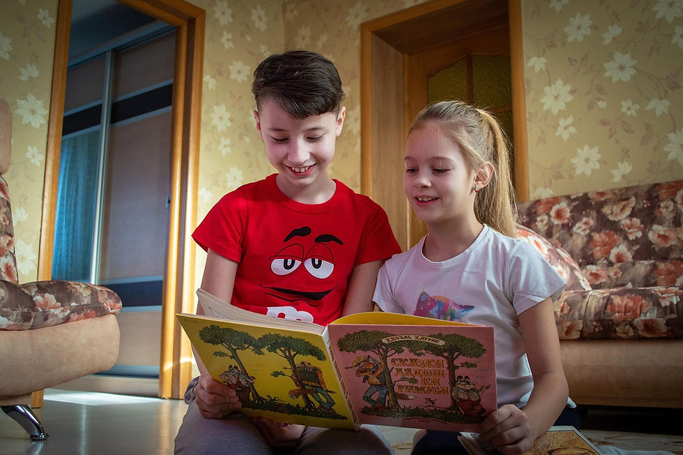 Two smiling children read a book together in a cozy, floral-patterned room. The boy wears a red shirt and the girl a white one.