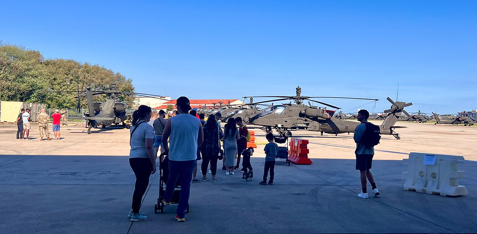 Crowd observes military helicopters on an airfield under clear blue skies. Families and individuals in casual attire engage with the scene.