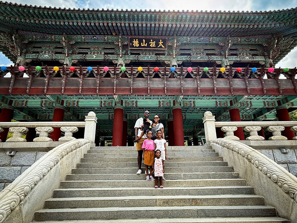 Family of six smiles on stone steps of a colorful, traditional Asian temple. Bright lanterns above, ornate pillars and roof details.