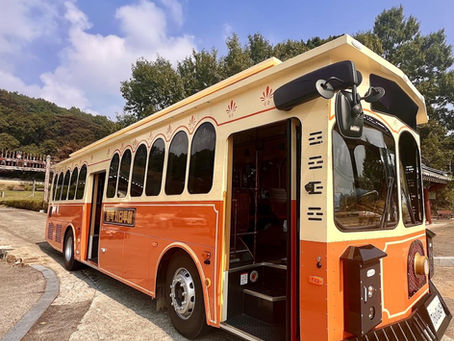 Orange vintage-style trolley bus parked on a sunny day. Open door, scenic background with trees and clear blue sky. Text in foreign script.