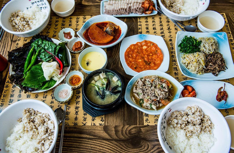 Korean meal spread with rice, soup, kimchi, and side dishes on a wooden table with Korean script. Vibrant and diverse food display.
