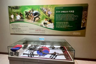 Display case with Korean flag and artifacts under glass. Green informational board with text and photos above, set in a museum-like environment.