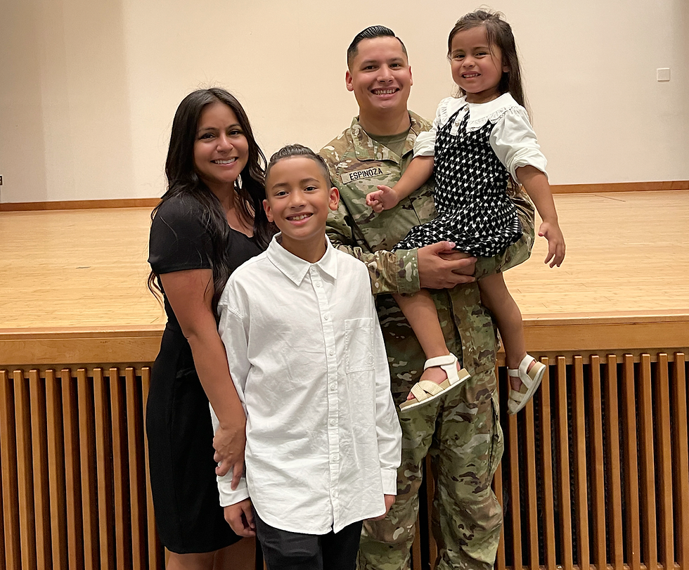 A smiling family poses indoors: a man in camouflage, woman in black dress, boy in white shirt, girl in black and white dress. Warm mood.