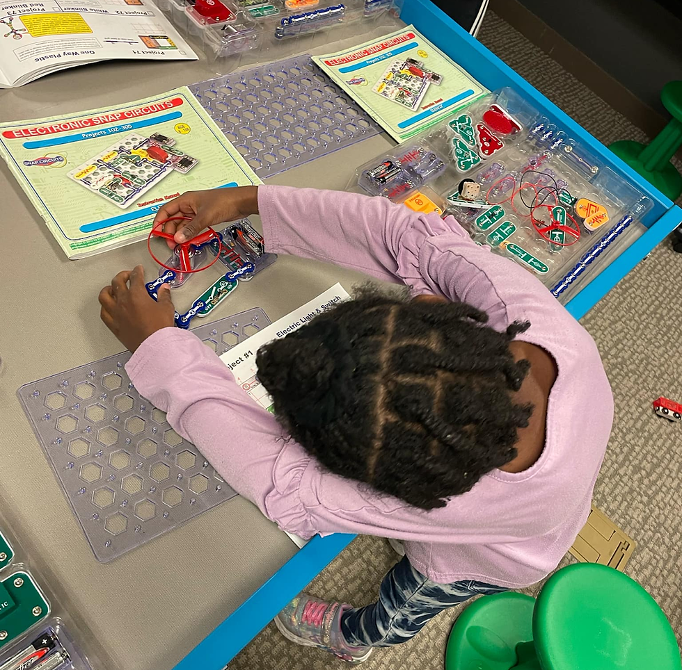 Child in purple shirt assembling a circuit kit on a table, with instructions and components around. Focused, learning activity in a classroom.