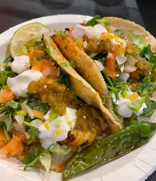 Two tacos filled with grilled meat, lettuce, tomatoes, sour cream, and salsa on a white plate. A lime slice and green chili pepper are on the side.