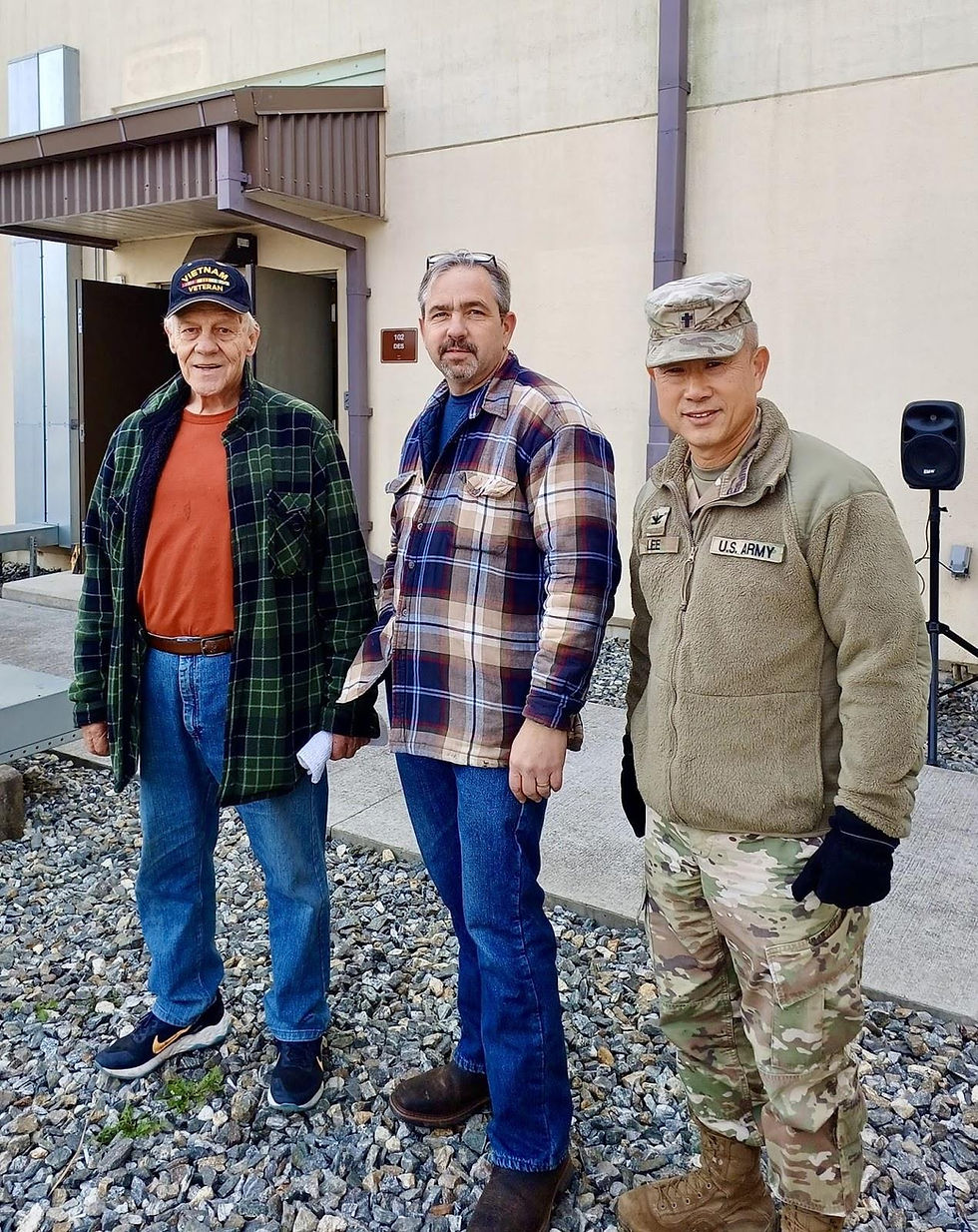 Three men stand outdoors: one in a veteran cap, another in plaid, and the third in Army attire. Gravel and a building are visible.