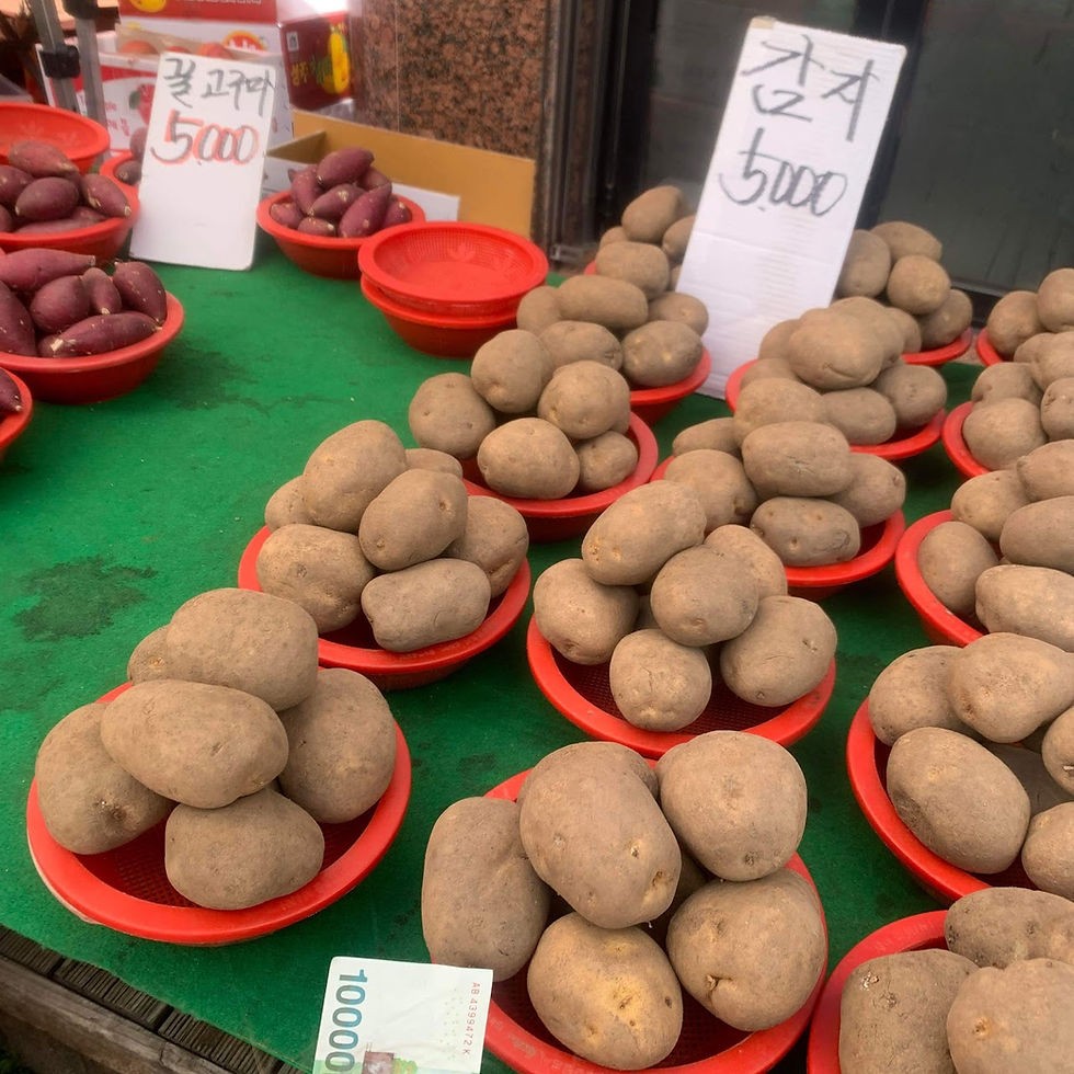 Potatoes in red baskets on a green mat at a market. Korean signs show prices as 5000. A currency note is visible. Bright and busy scene.